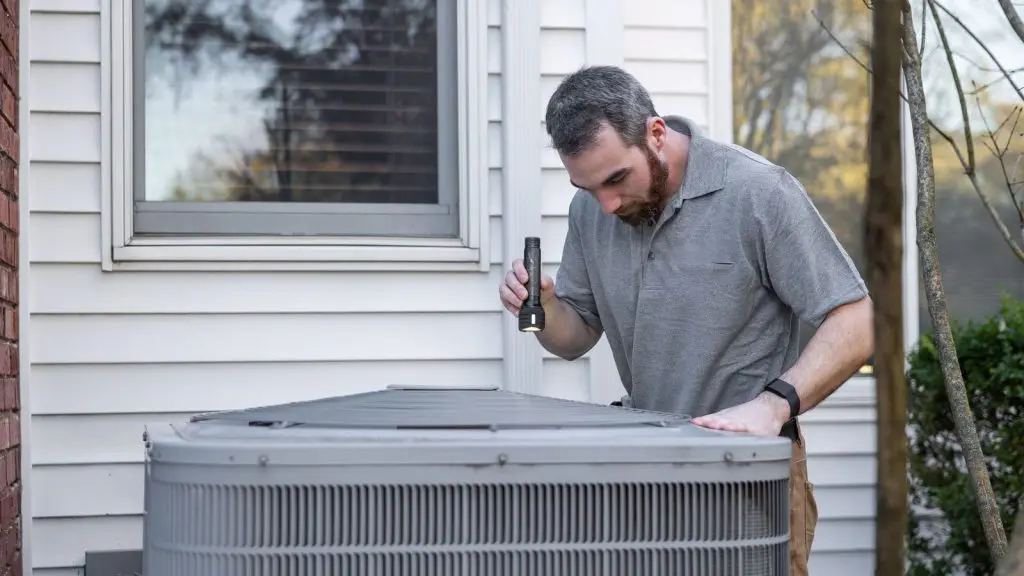 Technician with a flashlight inspecting an outdoor AC condenser, the work an HVAC marketing agency promotes