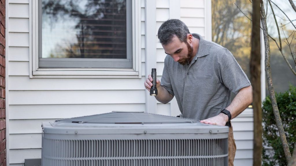 Technician with a flashlight inspecting an outdoor AC condenser, the work an HVAC marketing agency promotes